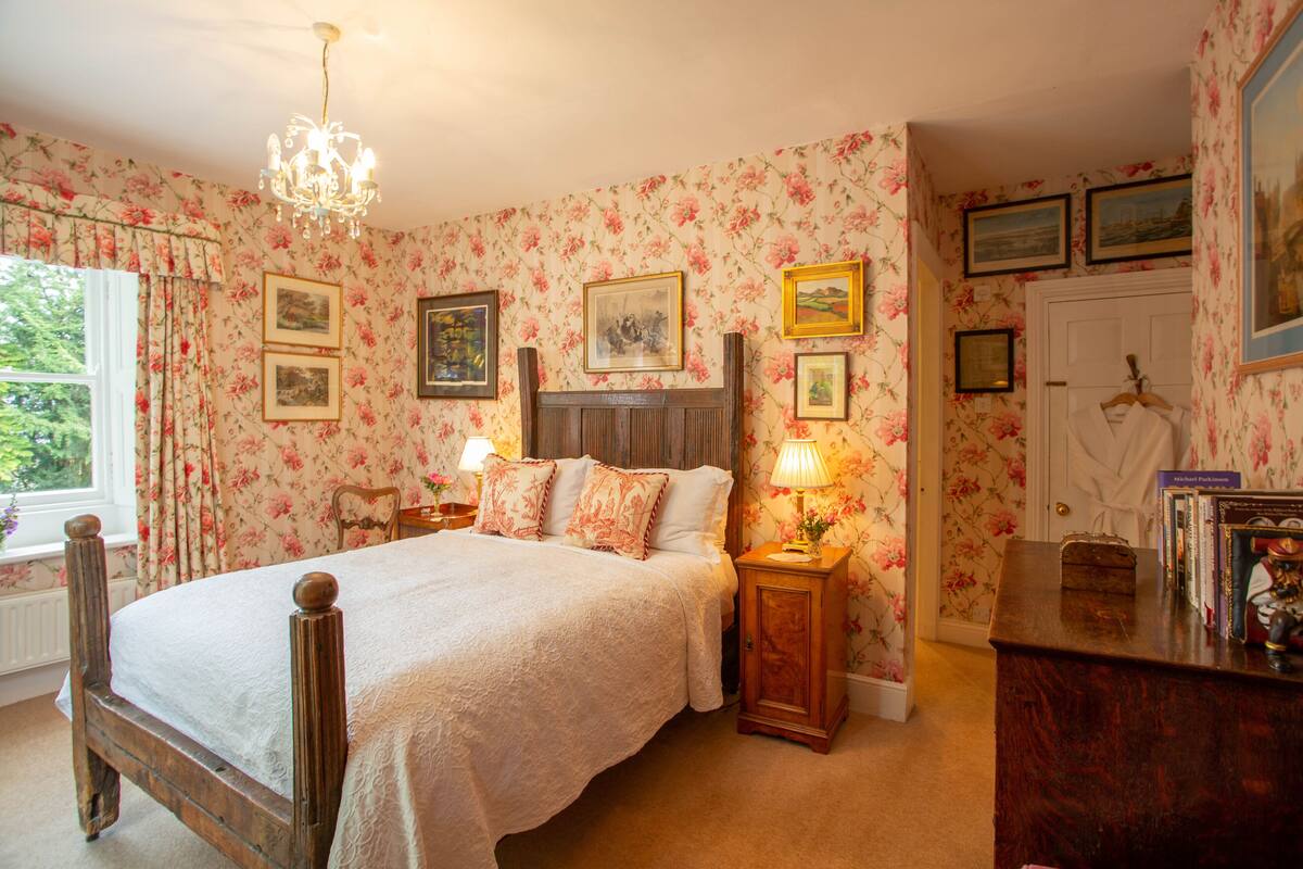 Ein Schlafzimmer mit floralem Wandpapier, einem hölzernen Bett, Nachttischen mit Lampen und gerahmten Bildern an den Wänden.