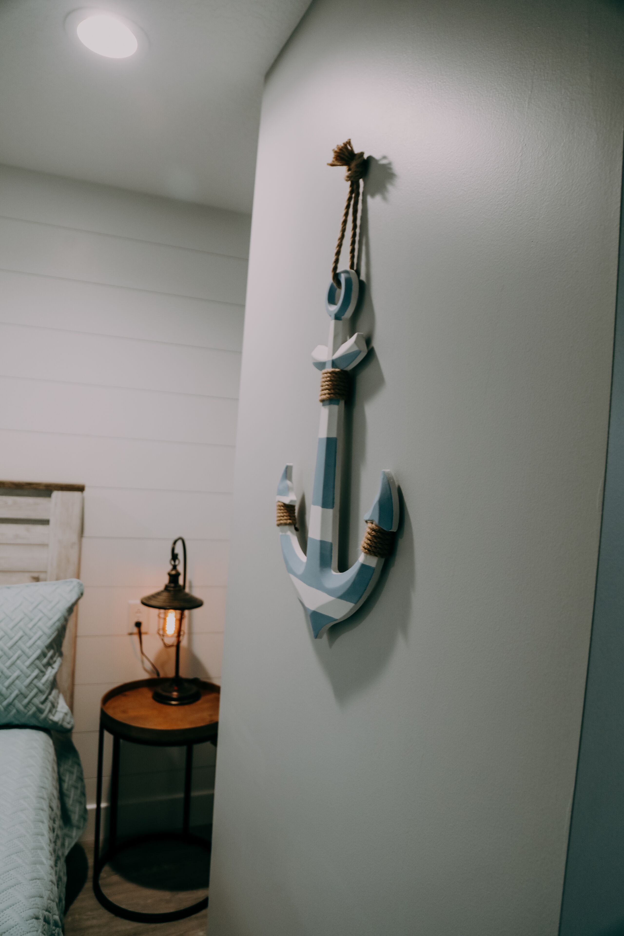 Een kamer met een bed, een klein rond tafeltje met een lamp en een decoratieve ankerhanger aan de muur.