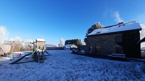 A snow-covered backyard with a wooden play structure, a stone house, and a clear blue sky.