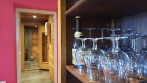 A room with wooden walls and a door leading to another area. A shelf holds several wine bottles and glasses.