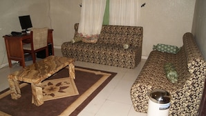 A living room with a patterned sofa, a wooden coffee table, a small table with a computer, and a white radiator. - Room-supreme Place Hotel - Guest Room (Abuja)