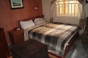 A bedroom with a bed, a nightstand, a wooden dresser, a bench, and a window with curtains.