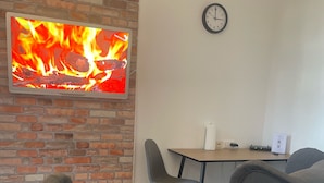 Un salon avec un mur en briques, un téléviseur à écran plat diffusant une image de feu de cheminée, deux fauteuils gris, une petite table blanche avec une télécommande et un pot, et une horloge au mur.
