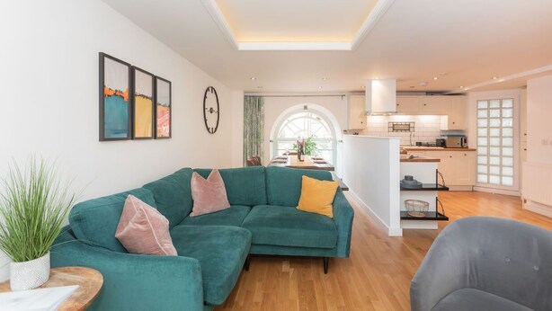 A modern living room with a sectional sofa, a dining area with a large window, and a kitchen in the background. - Kensington Chapel Apartments in Bath  (Bath)