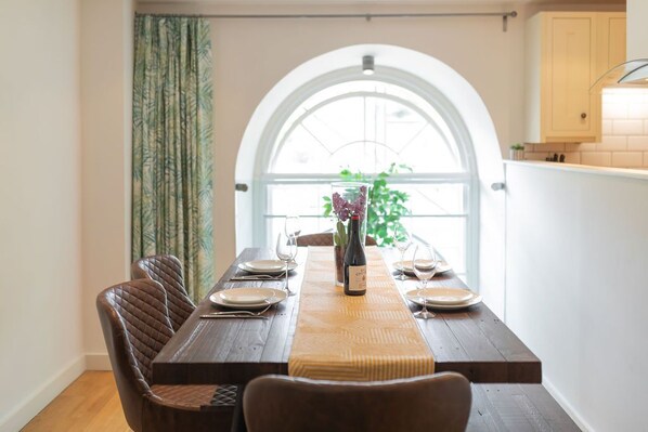 A dining area with a wooden table set for four, a vase with flowers, and a view through an arched window. - Kensington Chapel Apartments in Bath (Bath)