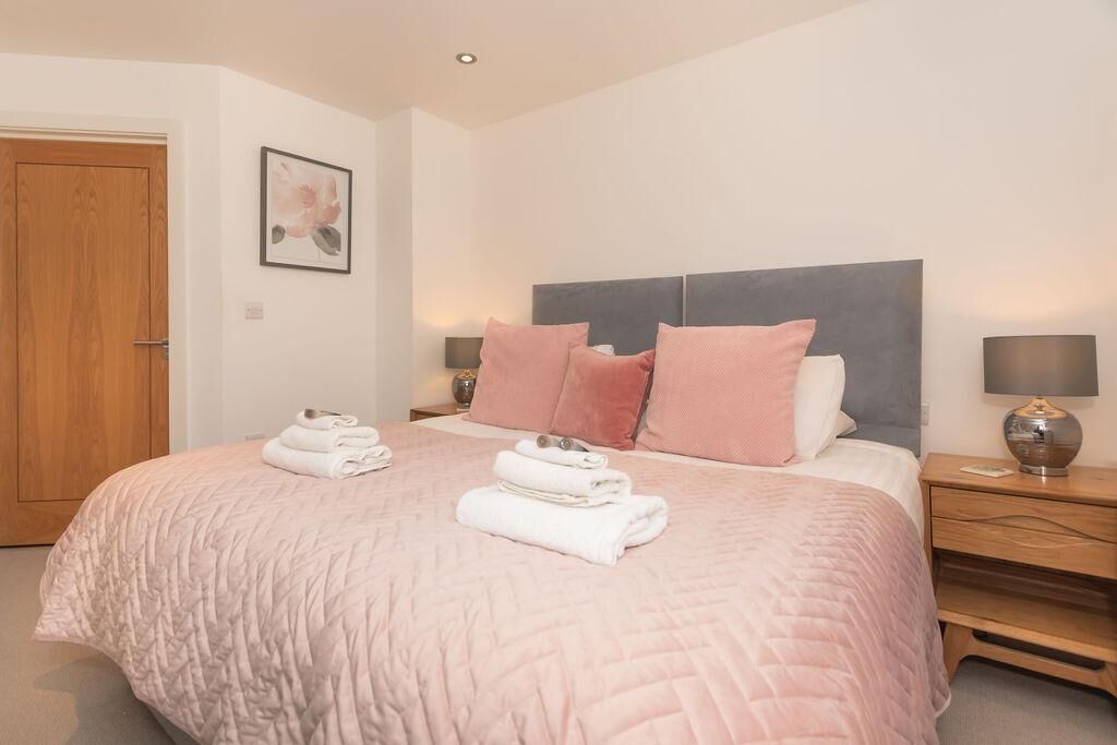 A neatly made bed with pink bedding and pillows, a wooden door, a framed picture on the wall, and a bedside table with a lamp.