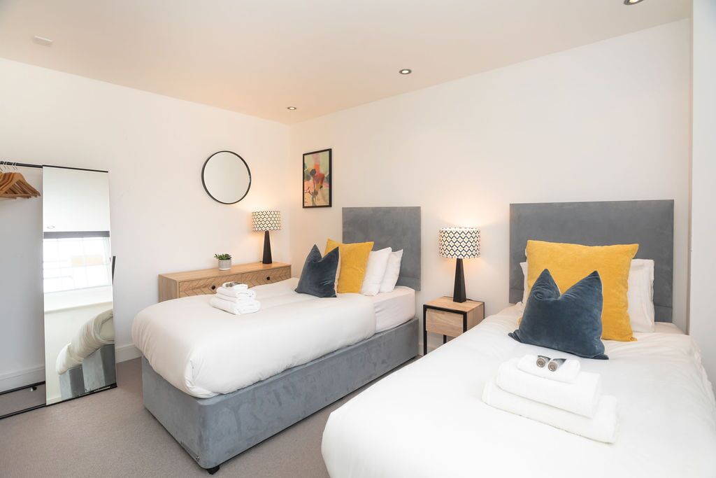 A modern hotel room with two beds, a large mirror, and a small side table with a lamp.