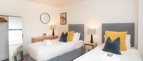 A modern hotel room with two beds, a large mirror, and a small side table with a lamp.