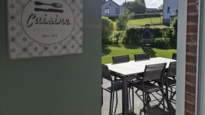 Un patio al aire libre con una mesa y sillas, un letrero colgante con la palabra 'Cuisine' y una fecha, y vistas de una zona residencial con casas y vegetación.