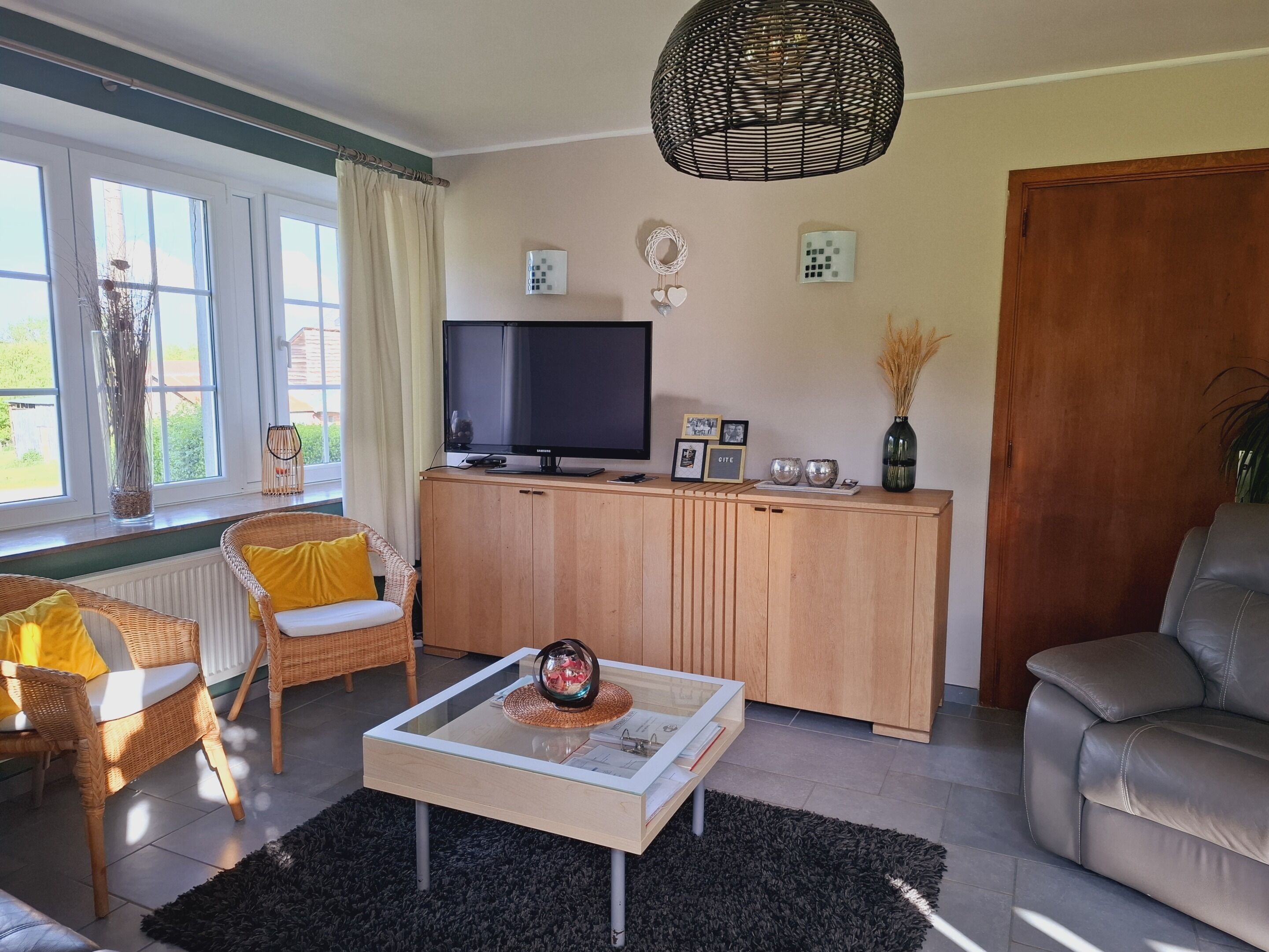 Een moderne woonkamer met een televisie, rieten meubels, een glazen salontafel en een groot raam met gordijnen.