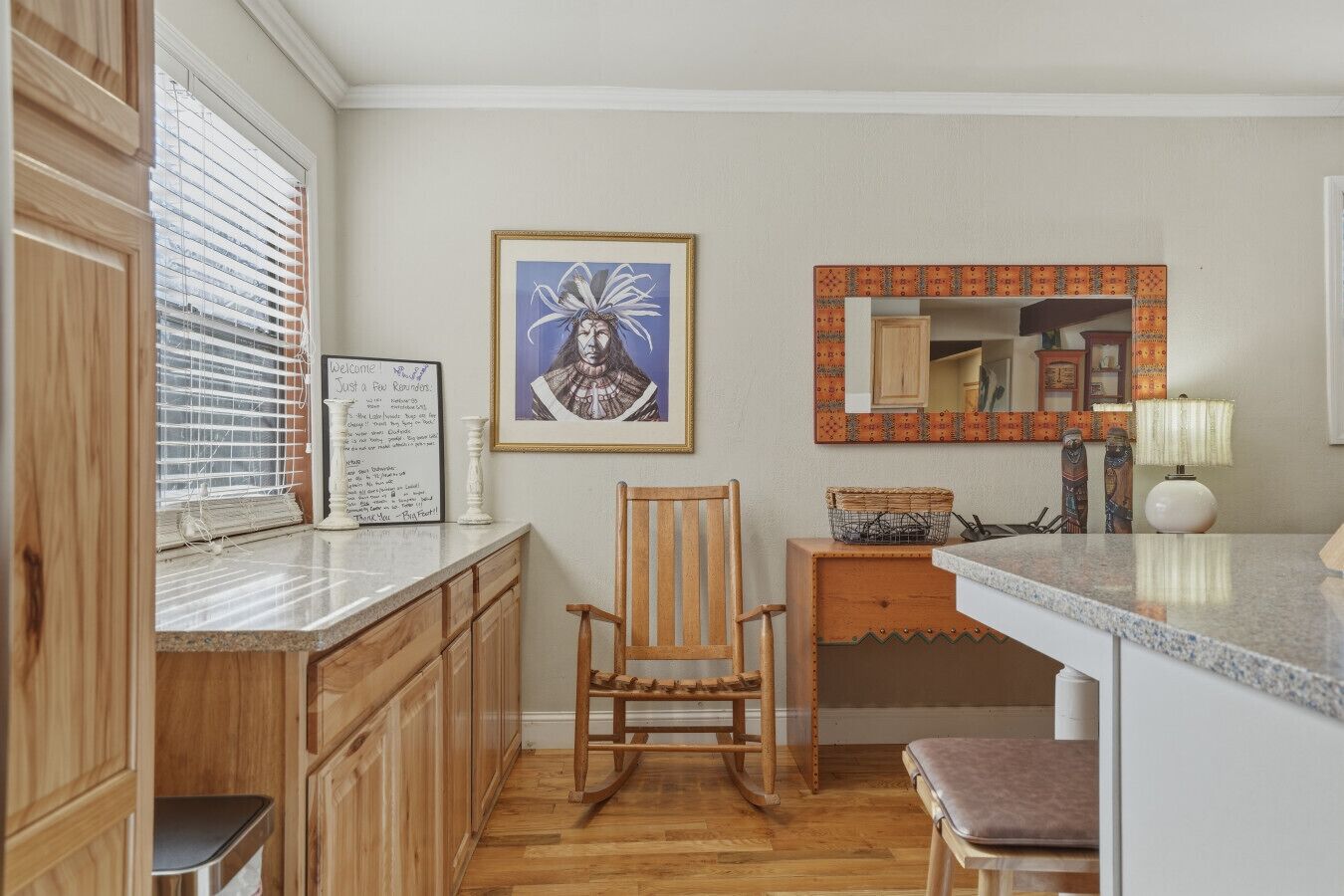 A kitchen with wooden cabinets, a rocking chair, a framed artwork, and a mirror with a patterned frame.