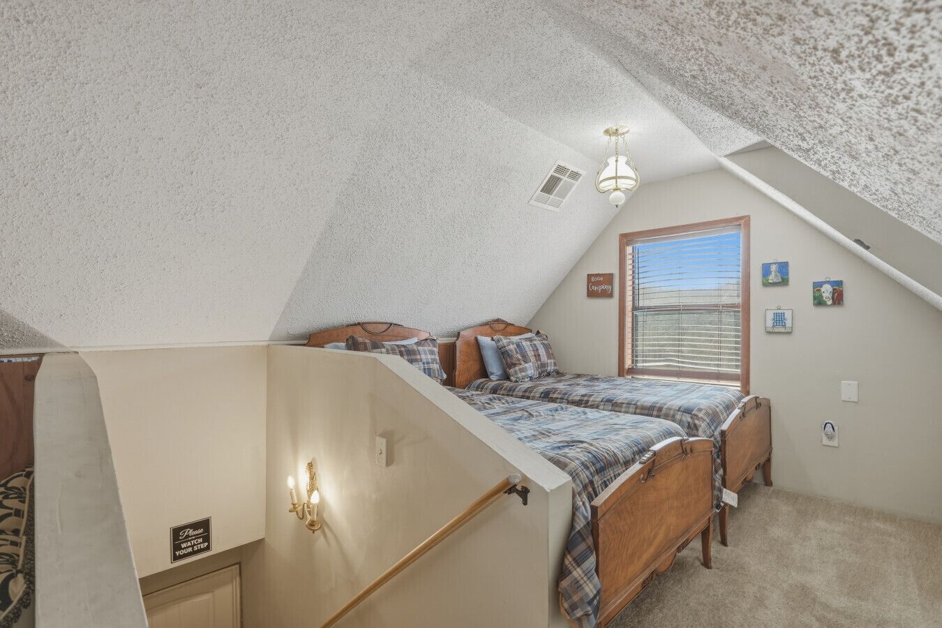 Une chambre avec un plafond en pente, un lit recouvert d’une couette à carreaux, une commode en bois et une fenêtre avec des stores.