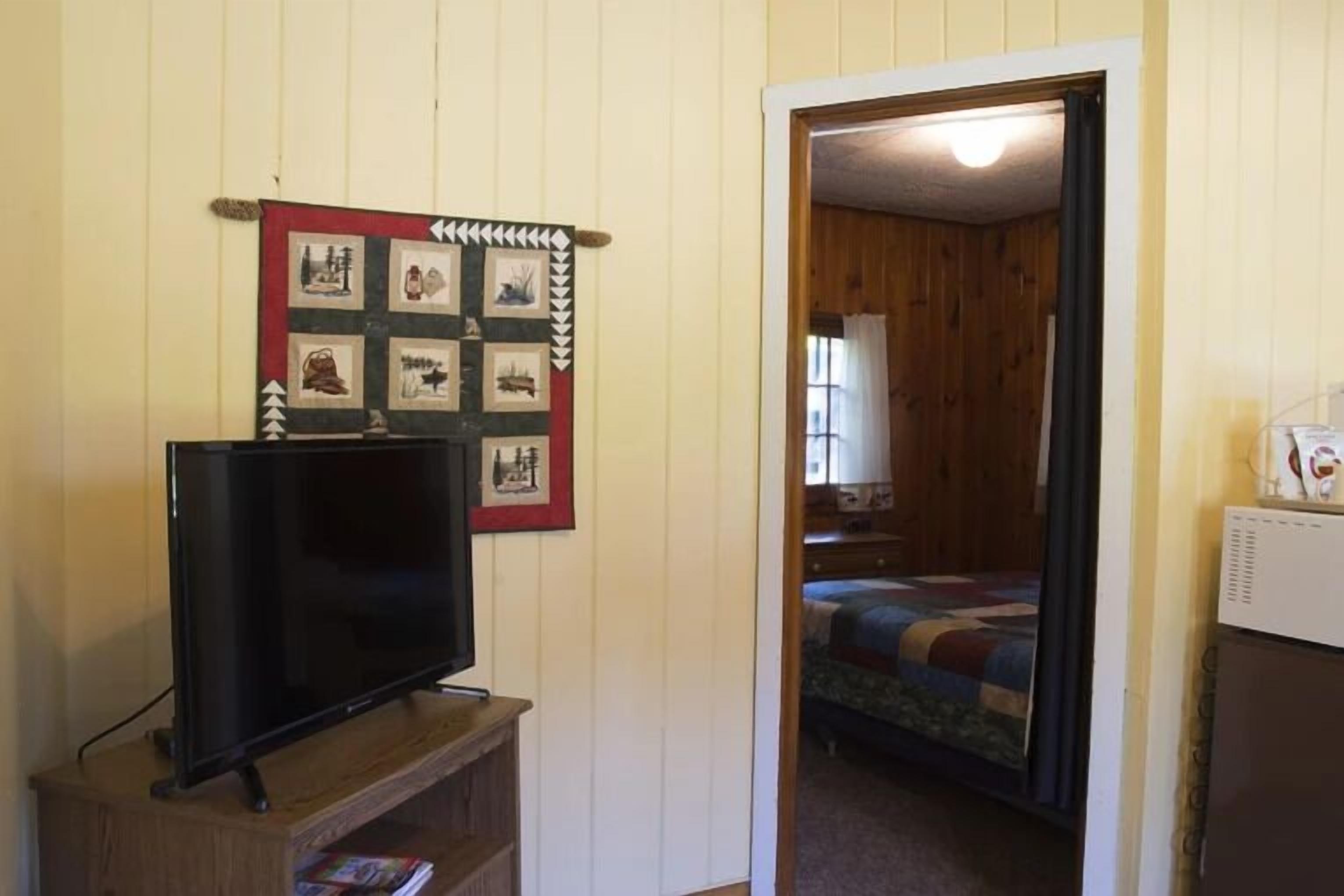 Une pièce avec un téléviseur posé sur un meuble en bois, une courtepointe ornée de divers motifs accrochée au mur, et un lit recouvert d’une couverture colorée.