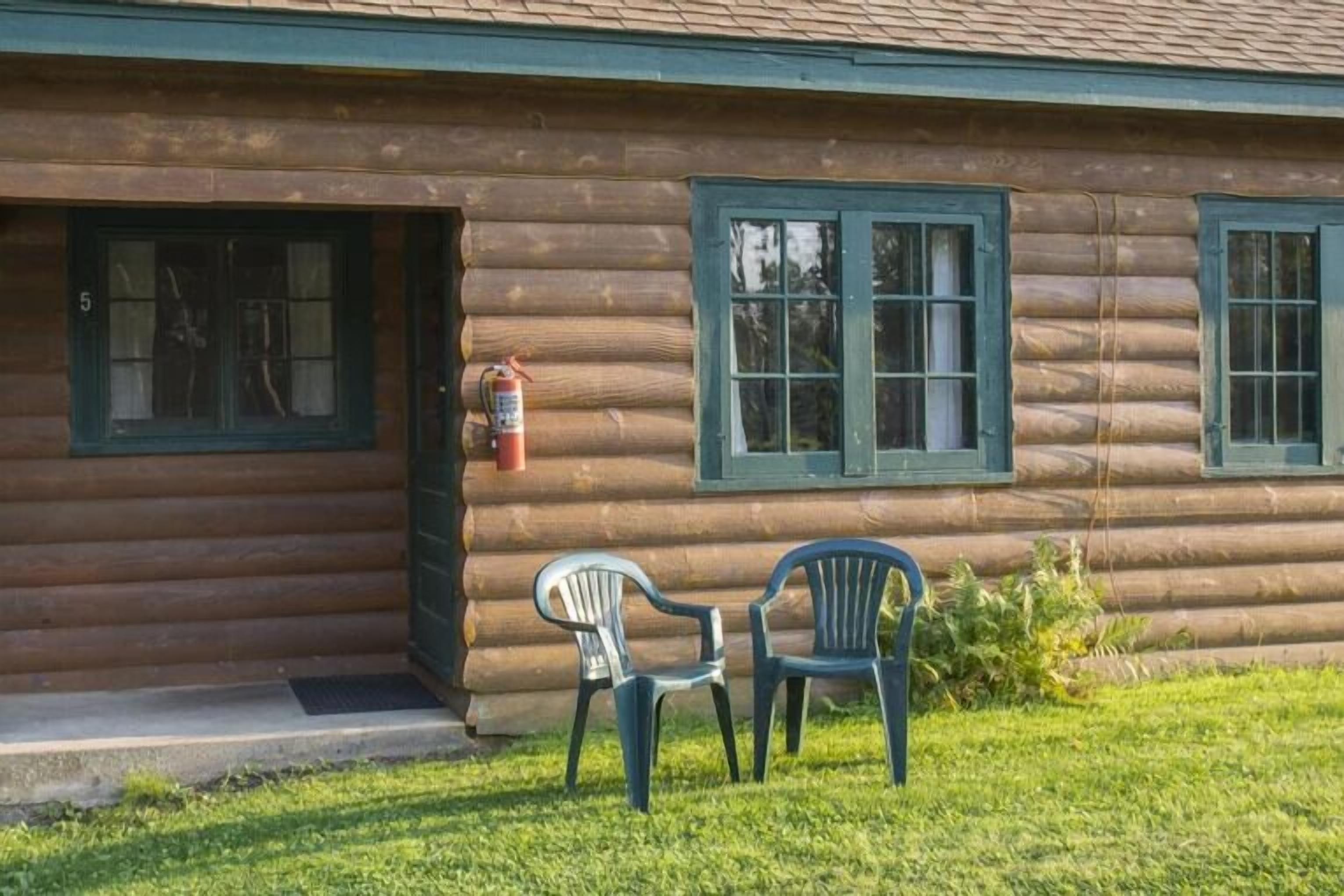 A log cabin with two windows, a fire extinguisher, and two chairs outside.