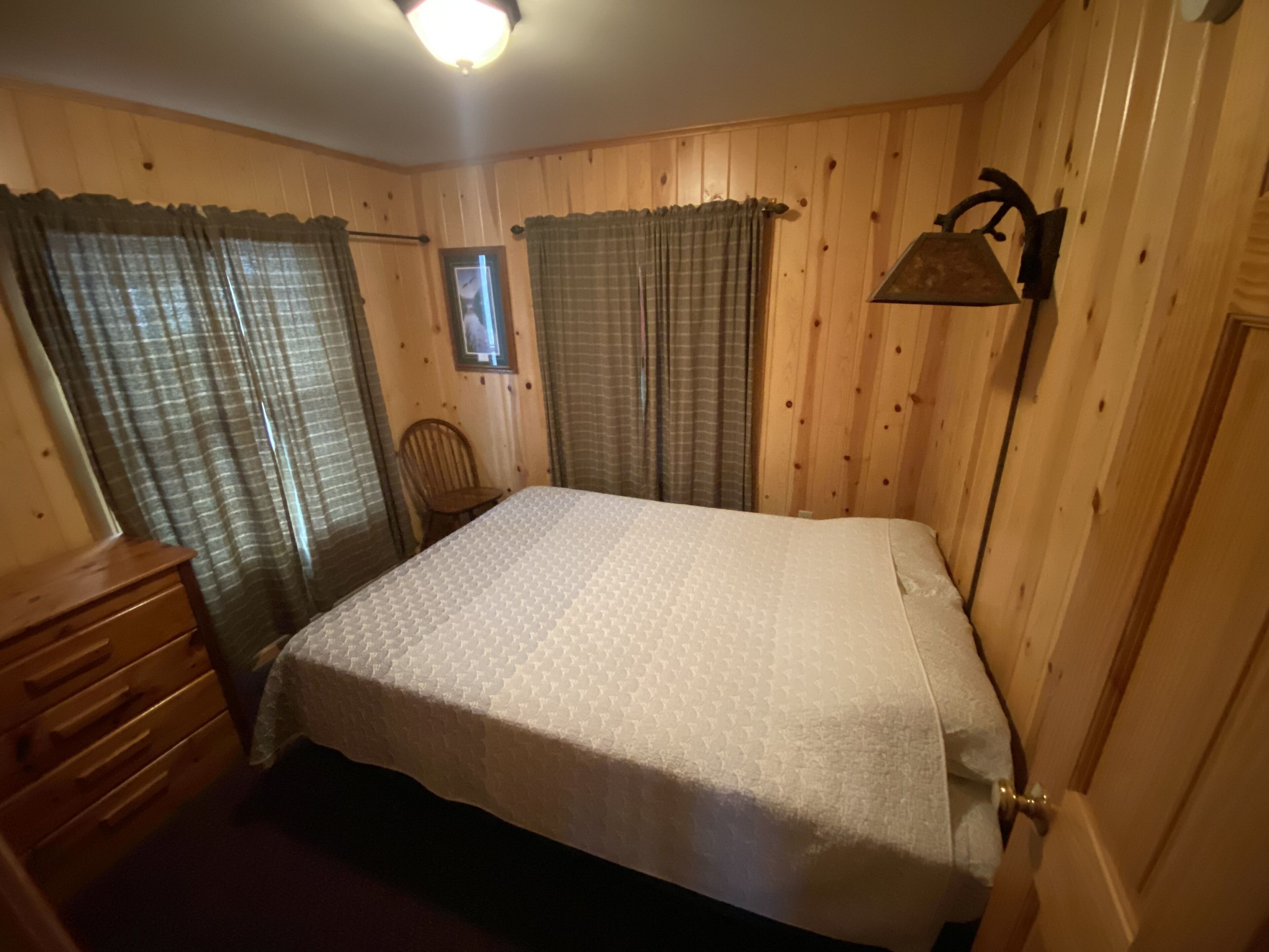 Ein Zimmer mit Holzwandverkleidung, einem Bett mit weißer Steudecke, einem Holzschrank und einer Wandlampe.