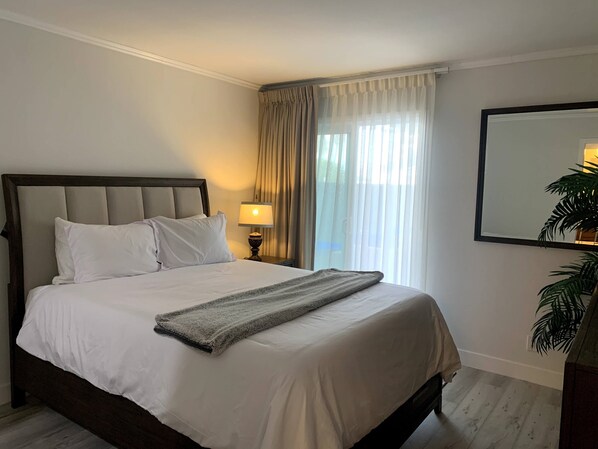 A neatly made bed with white linens and a grey throw blanket, a bedside lamp, and a large mirror on the wall.