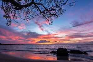 A beach scene with a sunset, calm sea, and rocky outcrops.