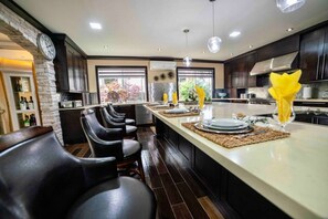 A modern kitchen with dark wood cabinets, a central island with bar stools, and a view of the outdoors through large windows. - Orchids & Palms Villa, Chef, Pool-Montego Bay! (Montego Bay)