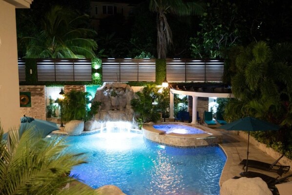 A nighttime pool area with a waterfall feature, lounge chairs, and a covered seating area.