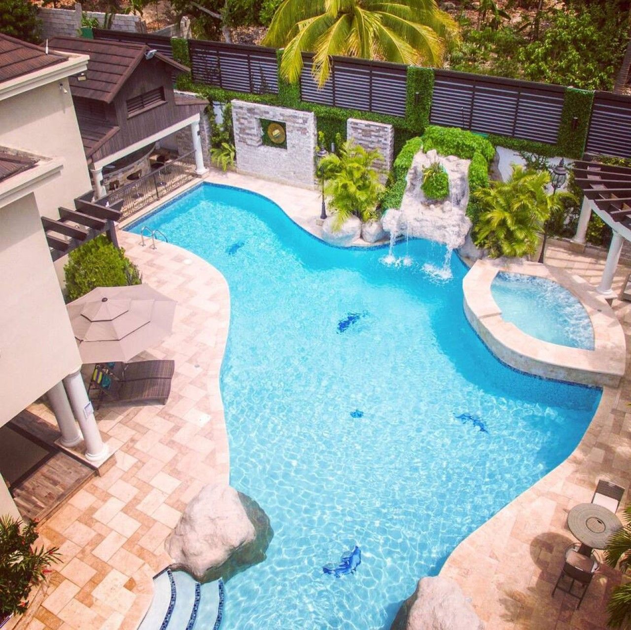 A swimming pool with a waterfall feature, surrounded by a landscaped garden and a building with a balcony.