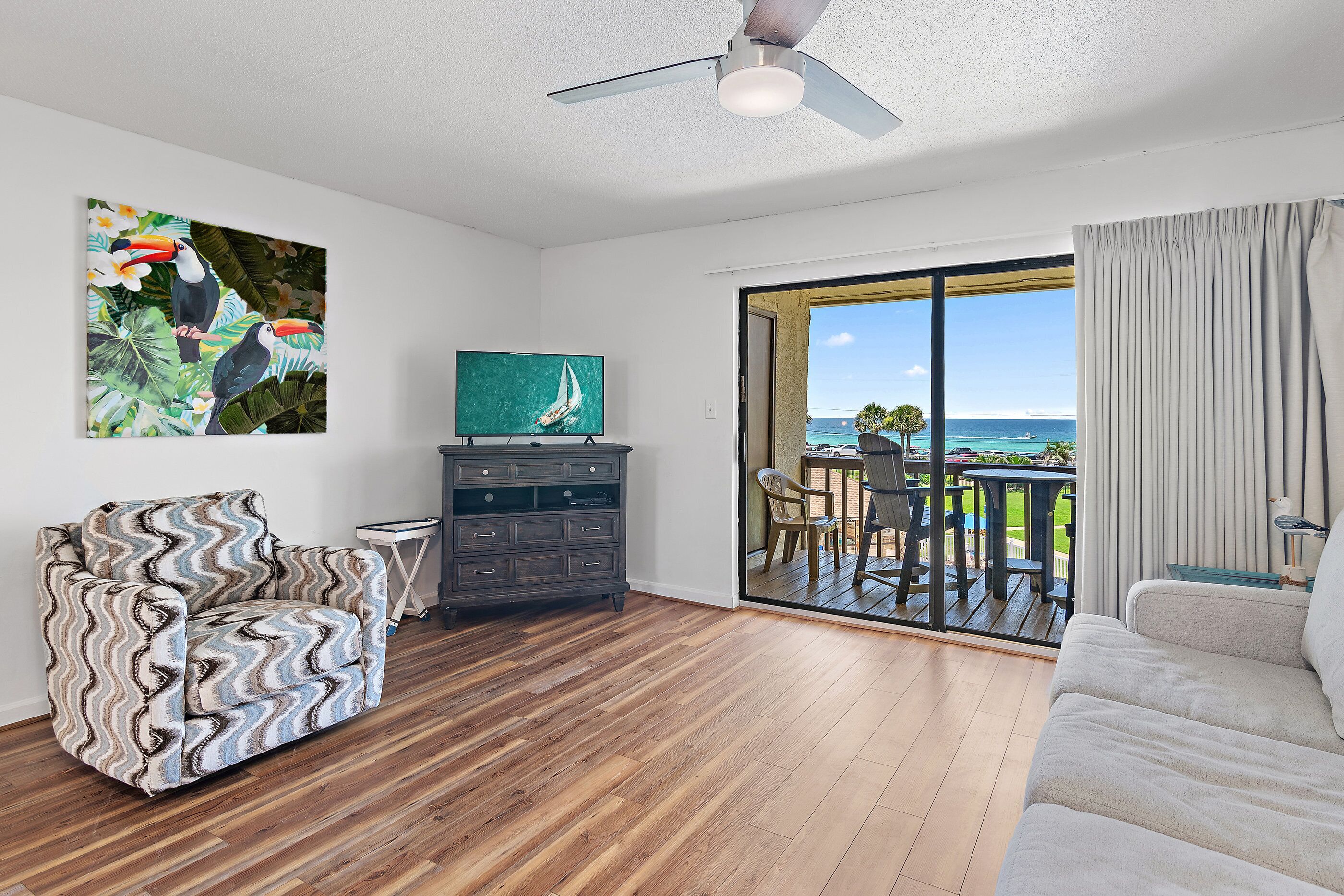 A living room with a wooden floor, a wavy-patterned armchair, a dark wooden cabinet, a television, and a view of the ocean through a sliding glass door.