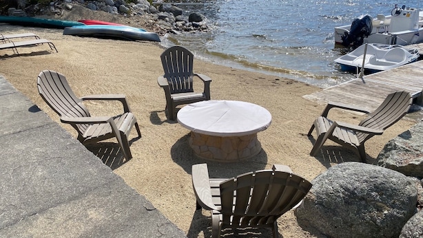 Una playa con sillas Adirondack y una mesa de piedra, rodeada de rocas y un bote fondeado cerca.