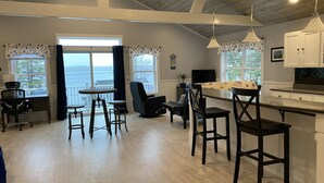 Amplia sala de estar con un gran ventanal con vista a un cuerpo de agua, comedor con sillas de madera y una mesa, y cocina con armarios blancos e iluminación colgante.