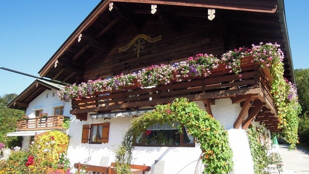 A traditional wooden chalet with a balcony, surrounded by vibrant flowers and a clear blue sky.