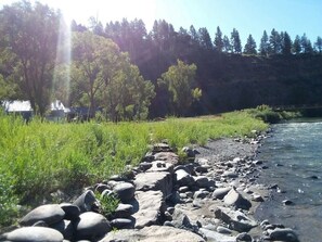 A riverbank with rocks and grass, a bridge in the background, and trees on a hillside. - Magical Riverside Retreat on 450 Private Acres (Pagosa Springs)