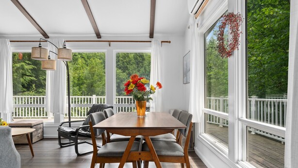 Ein überdachter Essbereich im Freien mit einem Holztisch und Stühlen, einer Blumenvase und Blick auf eine weiße Terrasse und Grünflächen.