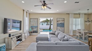 A modern living room with a large grey sectional sofa, a flat-screen TV, a ceiling fan, and a dining area with a chandelier. - Heated Pool + Games | Golf Cart | Near Beach (Jacksonville Beach)