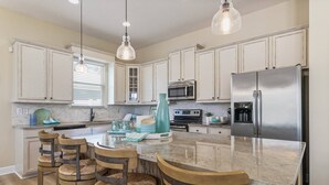 A kitchen with white cabinets, a stainless steel refrigerator, a large island with bar stools, and pendant lighting. - Heated Pool + Games | Golf Cart | Near Beach (Jacksonville Beach)