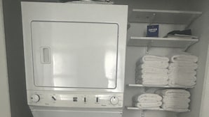 A compact laundry room with a front-loading washer and dryer, a shelf with folded towels, and a small shelf with a dryer manual.