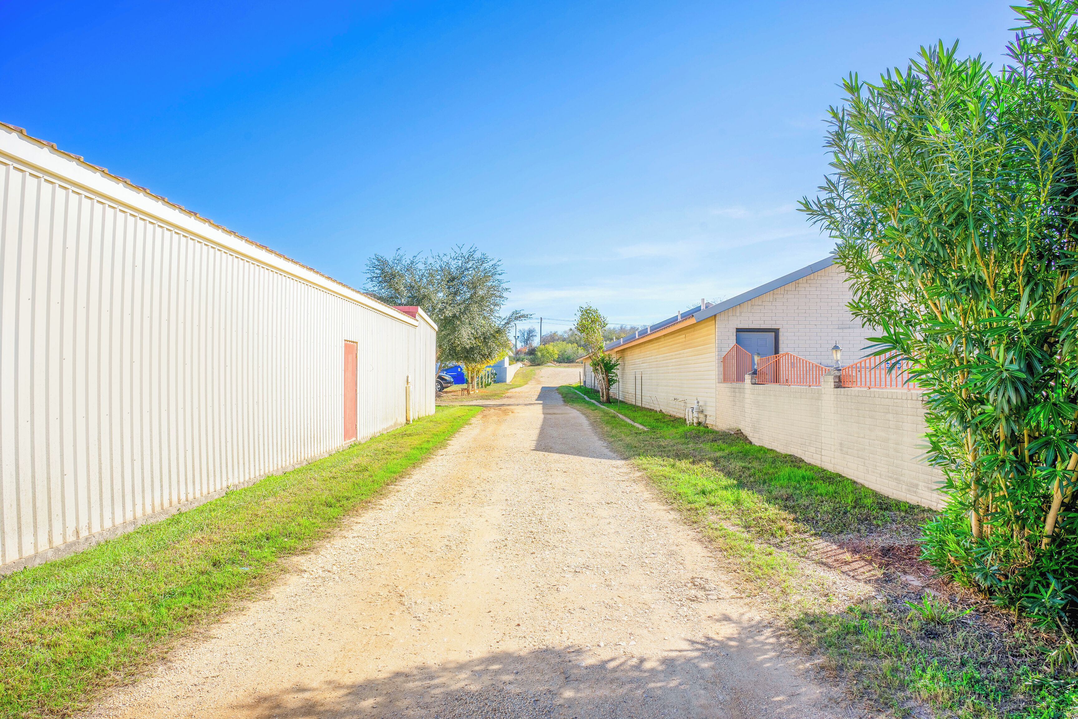 A narrow gravel path flanked by buildings with corrugated metal walls and a small tree on the right.