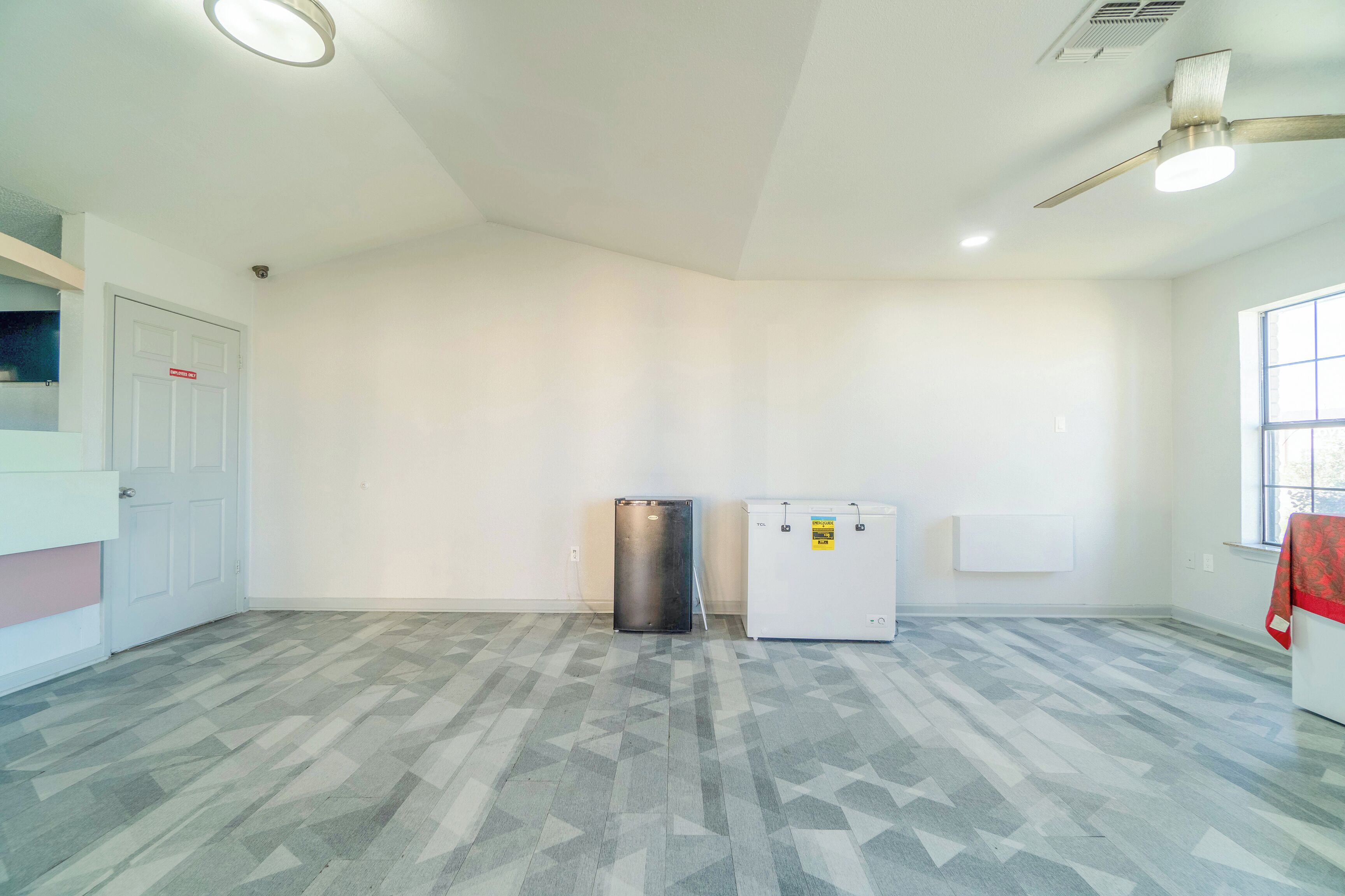 A spacious room with a geometric patterned floor, a white door, a small refrigerator, and a window with blinds.