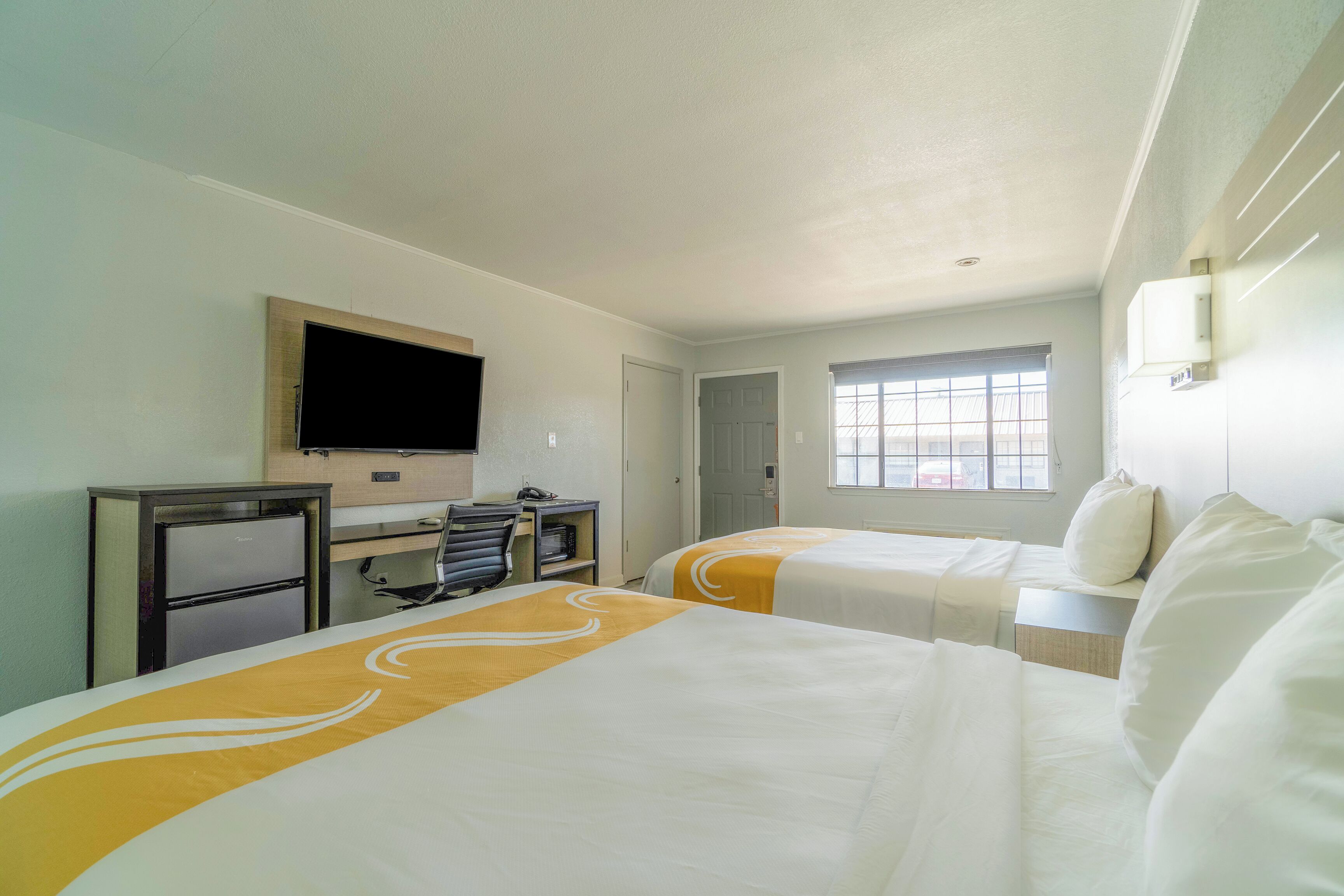 A hotel room with two beds, a desk with a computer, a TV, and a window with blinds.
