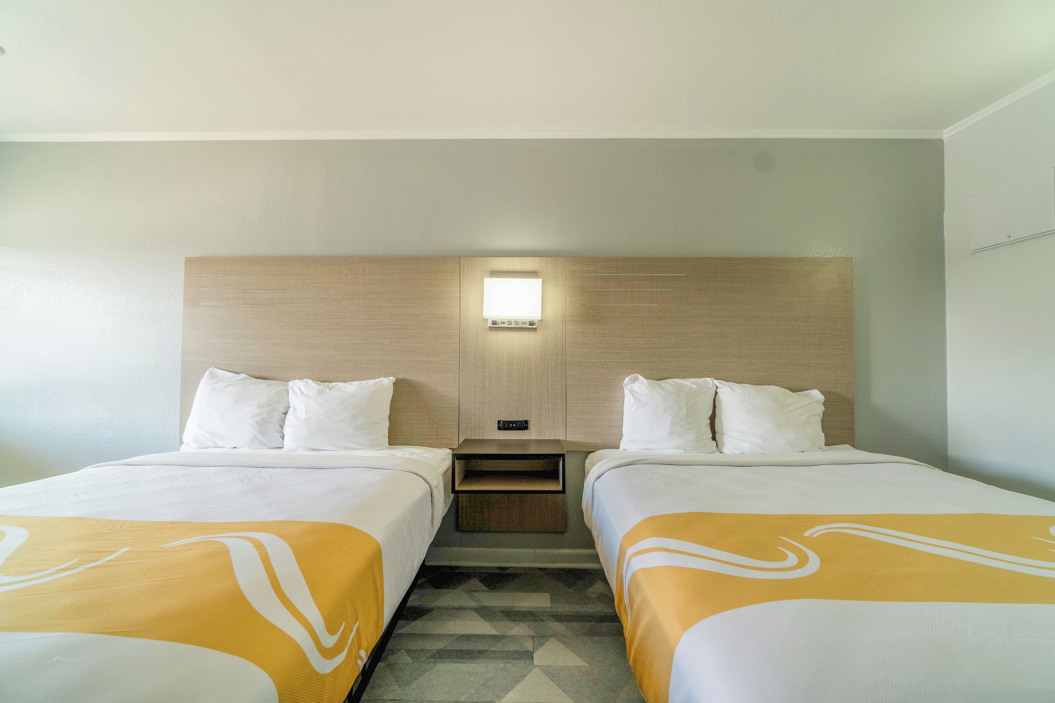 A hotel room with two single beds, a nightstand, a wall-mounted light, and a small shelf.