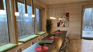 Ein langer Holztisch mit roten Platzsets, gepolsterten grünen Bänken und Metallstühlen in einem Raum mit Holzwänden und großen Fenstern.