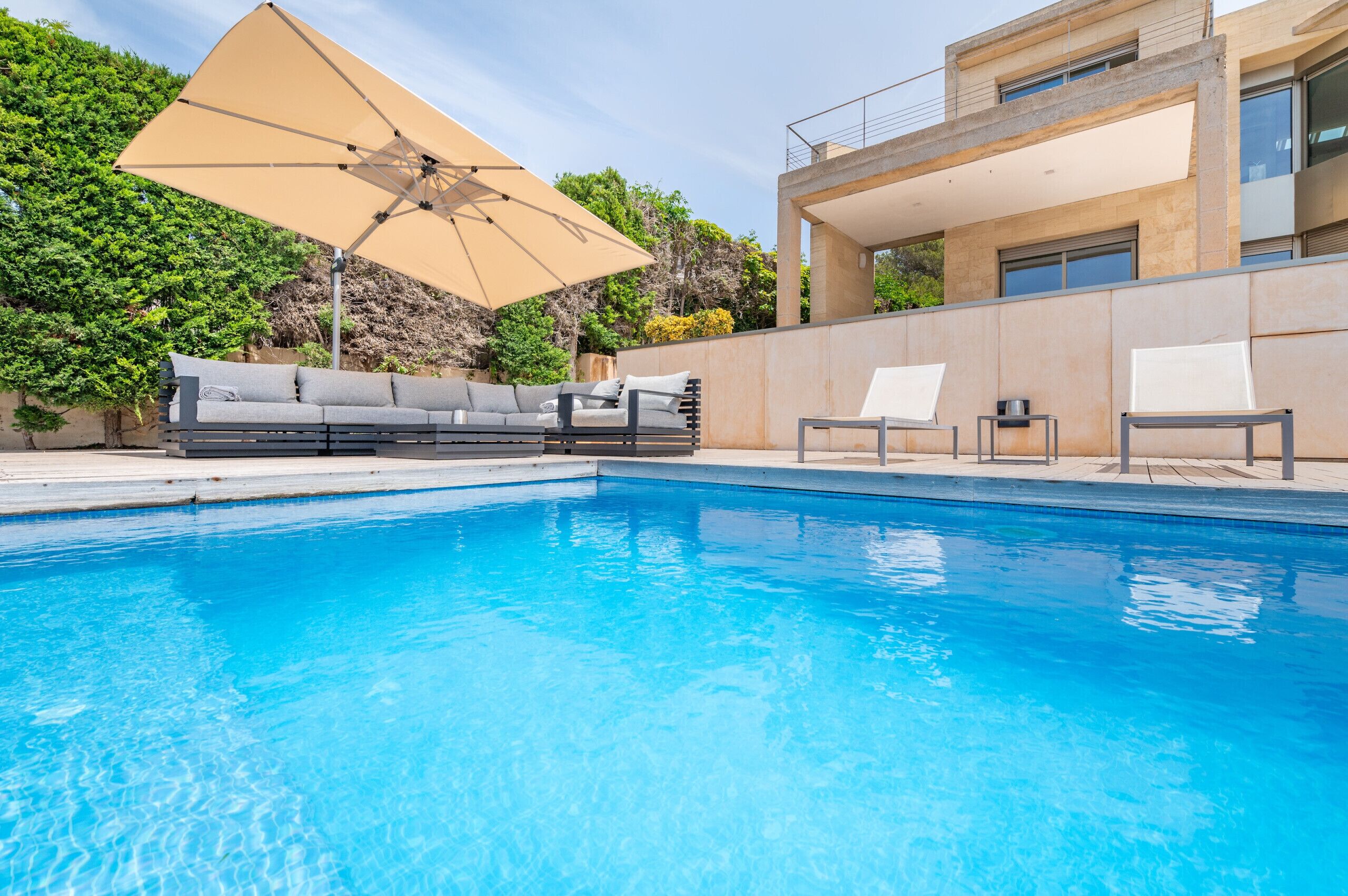 En swimmingpool med ett beige parasoll, en grå soffa och två solstolar.