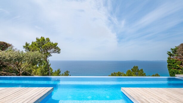 A clear view of a swimming pool with a wooden deck, surrounded by greenery and overlooking the sea. - Villa Beachfront - Herce Property by Priority (Llucmajor)