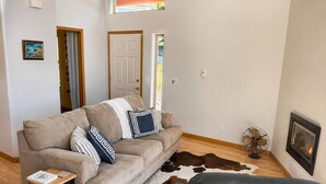 A living room with a sofa, a small table, a fan, a leather ottoman, a cowhide rug, a framed picture, and a door.
