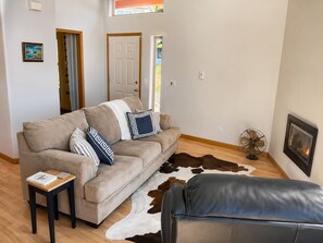 A living room with a sofa, a small table, a fan, a leather ottoman, a cowhide rug, a framed picture, and a door.