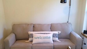 A living room with a sofa, a 'welcome' pillow, a lamp, and a coffee table with books.