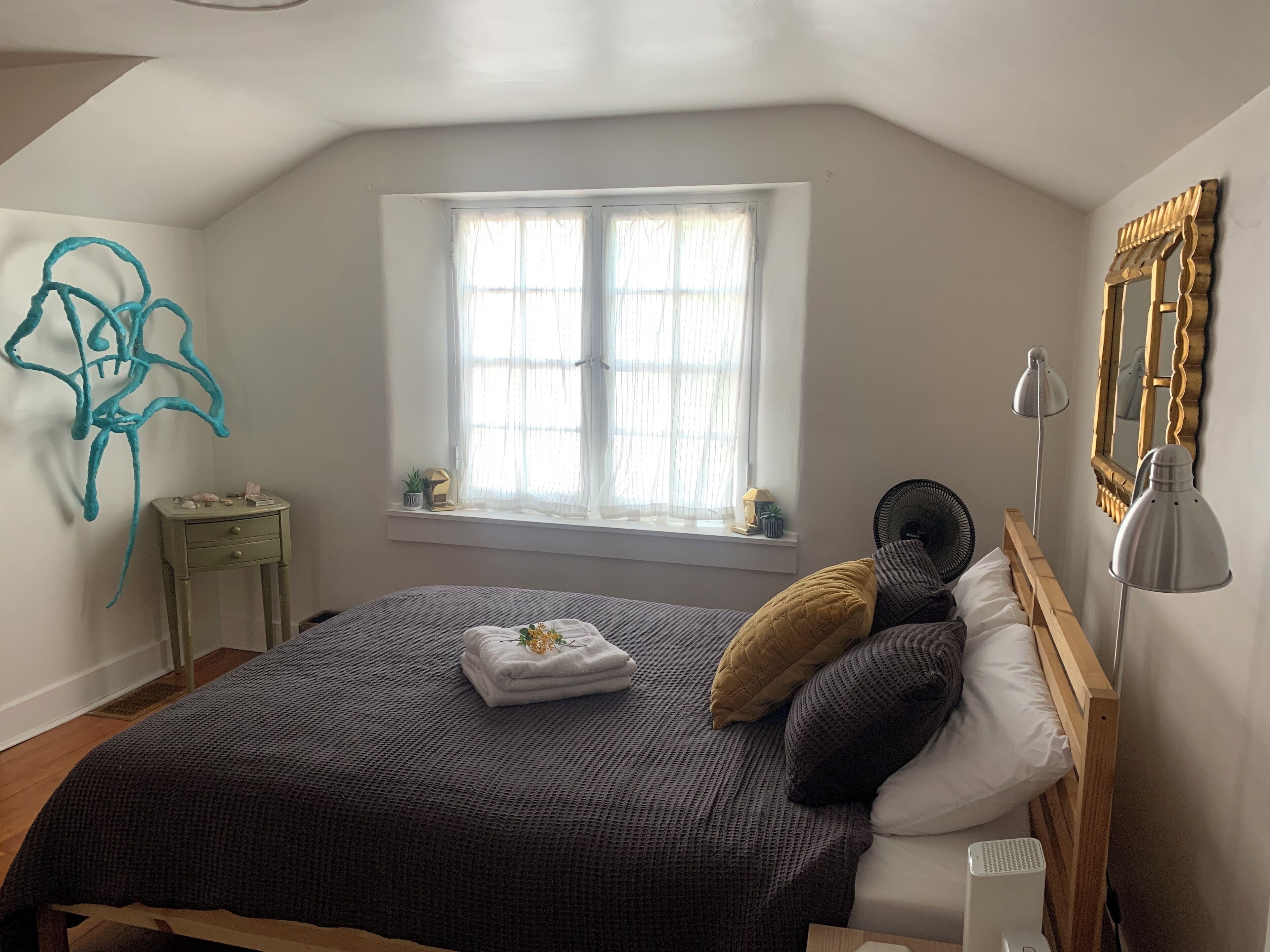A bedroom with a large bed, a window with sheer curtains, a bedside table with a lamp, and a wall-mounted mirror.