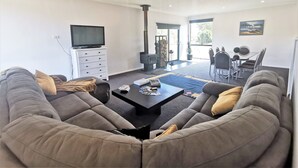 A spacious living room with a large gray sofa, a black coffee table, and a television on a white cabinet. There is a fireplace, a dining area with a table and chairs, and a large window providing natural light. - A Yingina Getaway, Home of Brown Trout (Miena)