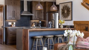 Een moderne keuken met donkerhouten kastjes, een centraal eiland met krukken, roestvrijstalen apparatuur, hanglampen en een decoratief wandobject.