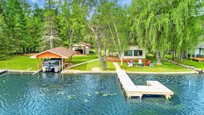 A clear swimming pool with a dock, a covered boat house, and a small cabin with outdoor seating. - Clam Lake Waterfront Cottage (Bellaire)