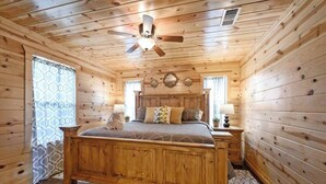 Un dormitorio de cabaña de madera con cama, dos mesitas de noche, ventilador de techo y ventana con cortinas estampadas.