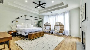 Une chambre à coucher avec un lit à baldaquin, deux fauteuils en osier, un banc en bois et une grande fenêtre ornée de rideaux légers.