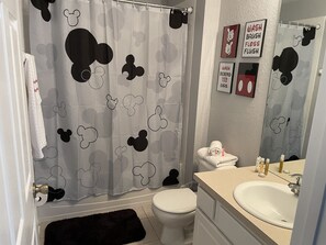 A bathroom with a Mickey Mouse themed shower curtain, a toilet, a sink with toiletries, and a mirror. - Disney Villa| Private Pool|5 mins to Disney|Resort amenities (Kissimmee)